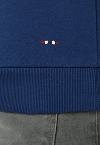 Suéter azul marino acanalado con un diseño corto bordado en rojo y blanco en la parte inferior. Material suave, debajo hay una tela texturizada gris.