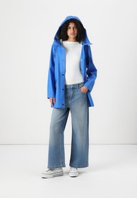 Imperméable bleu avec capuche, devant à boutons et cordons de serrage. Associé à un pantalon en denim à jambes larges et des sneakers blanches.