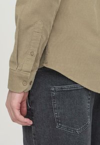 Un primo piano del braccio di una persona in una camicia di velluto a coste color khaki, con i bottoni visibili, accanto a jeans in denim scuro. L'attenzione è sulla texture del tessuto.