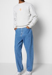 Sudadera gris con un gráfico naranja en el pecho, combinada con unos vaqueros de mezclilla azul de corte holgado y zapatillas blancas.