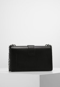 Černá obdélníková kožená kabelka s klopou a stříbrným řetízkovým popruhem, vystavená na bílém podstavci proti jednoduchému pozadí.