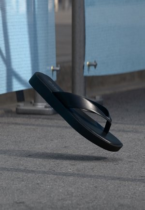 Black flip-flop sandal suspended mid-air above asphalt, with blue fabric panels and metal poles in the background.