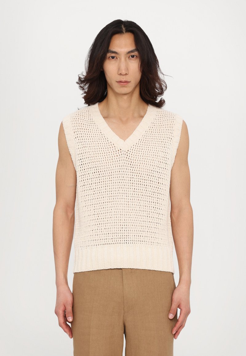 Gilet sans manches en crème clair avec un col en V, un dessin en mailles ouvertes et un ourlet côtelé. Associé à un pantalon beige.