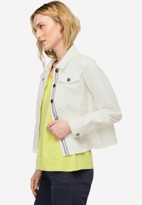 Weißer Denim-Jacke mit Druckknöpfen, Brusttaschen und einem kontrastierenden schwarzen Streifen an der Leiste, kombiniert mit einem leuchtend grünen Top.