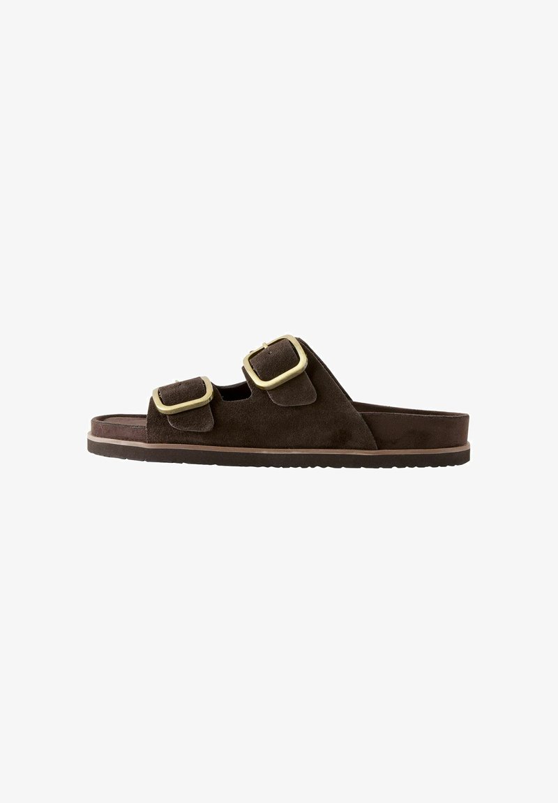 Bruine suède slip-on sandaal met twee verstelbare gouden gespen en een platte rubberen zool, getoond vanaf de zijkant op een witte achtergrond.
