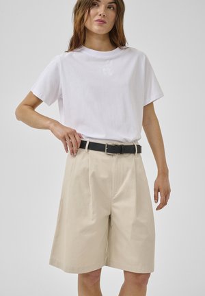 Nő fehér, rövid ujjú pólót visel, amelyet bézs színű térdig érő, bő szárú rövidnadrágba tűrt, fekete övvel, egyszínű háttér előtt áll.