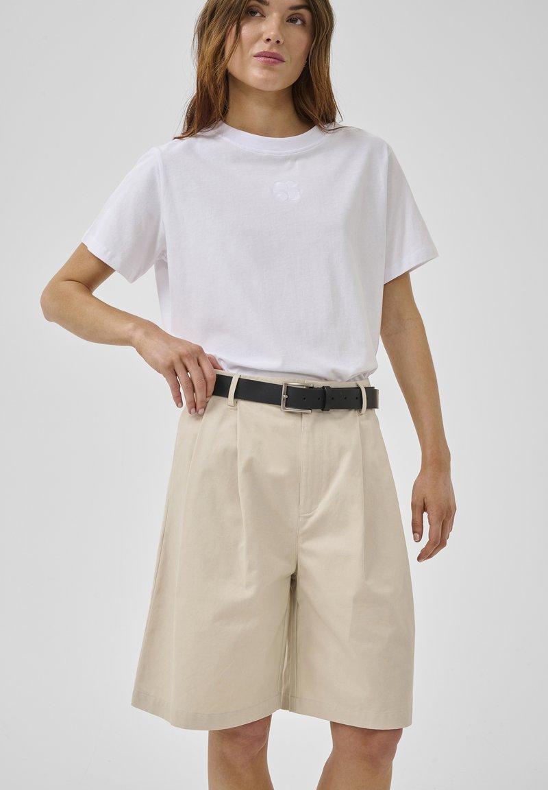 Femme portant un t-shirt blanc à manches courtes rentré dans un short beige large coupé aux genoux avec une ceinture noire, debout devant un fond uni.