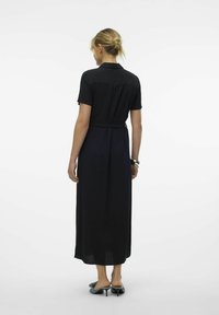Robe maxi noire à manches courtes, avec col et détail de ceinture. Tissu plat avec une texture lisse. Portée avec des mules à talons bleus.