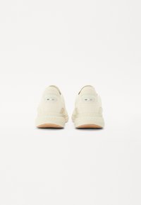 Light beige sneakers with a smooth texture, rounded shape, and rubber soles. Features a subtle logo detail on the heel.