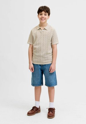 Jeune garçon debout, portant un polo beige en maille à manches courtes, un short en jean bleu, des chaussettes blanches et des chaussures en cuir marron, souriant à la caméra.