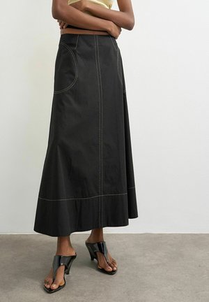 Femme portant une jupe midi noire avec des coutures contrastantes, associée à des sandales à talons noires à bout ouvert, debout les bras croisés.