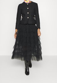 Černá texturovaná bunda se zlatými knoflíky, doplněná dlouhou vrstvenou černou tylovou sukní. Černé kozačky po kolena dokončují celý vzhled.