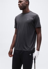 Άνδρας που φοράει σκούρο γκρι T-shirt Calvin Klein και μαύρο παντελόνι Adidas, κρατώντας ένα γκολφ κλαμπ με λευκό γάντι στο δεξί χέρι, στραμμένος προς τα αριστερά.