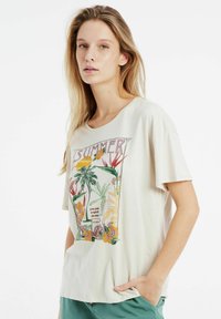 Camiseta de manga corta color crema con un gráfico tropical colorido y texto, corte relajado y textura de tela suave.