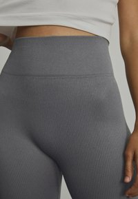 Szürke bordázott leggings, magas derékkal, sima textúrával. Klasszikus részletmegtekintés, amely bemutatja a derékrészt és a szövet részleteit.