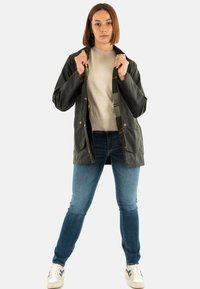 Vrouw met schouderlang haar, gekleed in een donkergroene jas over een beige trui, blauwe spijkerbroek en witte sneakers, staand tegen een witte achtergrond.