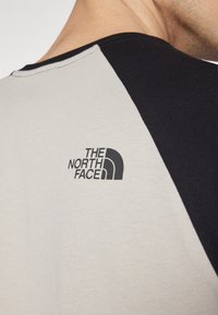 Camiseta de manga corta en gris claro con mangas raglán negras. Presenta un logotipo negro en la parte superior de la espalda. Fabricada con un tejido suave y transpirable.