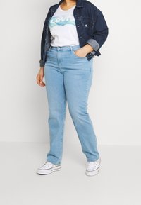 Giacca di denim blu sopra una maglietta bianca con una grafica colorata, abbinata a jeans straight-leg blu chiaro e sneakers bianche.
