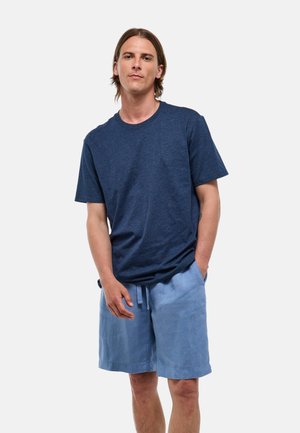 Jeune homme debout, une main dans la poche, portant un t-shirt bleu marine et un short bleu clair à cordon, sur un fond uni.