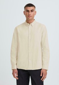 Camisa beige de manga larga hecha de tela suave, con cuello abotonado, botones en la parte delantera y un bordado sutil en el pecho.
