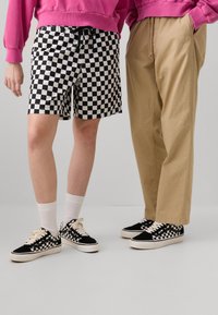Shorts de cuadros en blanco y negro con una cintura elástica, combinados con pantalones de pierna ancha en color beige. Ambos llevan zapatillas de skate negras con acentos de cuadros.