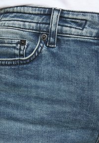 Primer plano de unos jeans de mezclilla azul claro, con una cinturilla con un botón y dos bolsillos frontales, mostrando la textura y la costura de la tela.