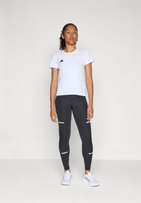 Maglietta atletica bianca a maniche corte con logo nero, abbinata a leggins scuri con strisce bianche. Modella che indossa scarpe da ginnastica bianche.