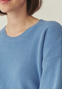 Pull en maille côtelée bleu clair avec un col rond et une coupe décontractée, présentant une texture visible et des détails de couture subtils sur les manches.