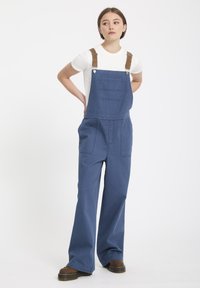 Blauwe spijkeroveralls met wijd uitlopende pijpen, schouderbanden met bruine accenten, een voorkantzak en een ontspannen pasvorm. Geraadpleegd met een witte T-shirt.