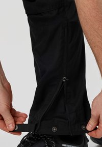 Χέρια που ρυθμίζουν το κλιπ και το ξεκούμπωτο μανίκι ενός μαύρου παντελονιού outdoor πάνω από μαύρο παπούτσι Swedemount.