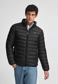 Sort pufferjakke med quiltet design, høj krave, og frontlynlås. Har sidelommer og et logo-accents på brystet.