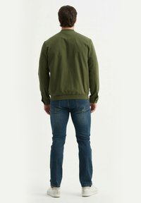 Veste bomber vert olive avec bords côtelés et poignets, associée à un jean en jean bleu et des baskets blanches, vue de l'arrière.