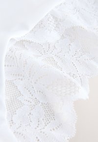 Détail de tissu en dentelle blanche avec des motifs floraux et des bords festonnés sur un fond de tissu blanc lisse.