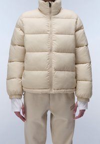 Beige pufferjacka med hög krage, horisontell sömnad, elastiska muddar och dragkedja framtill. Bärs över matchande beige byxor.