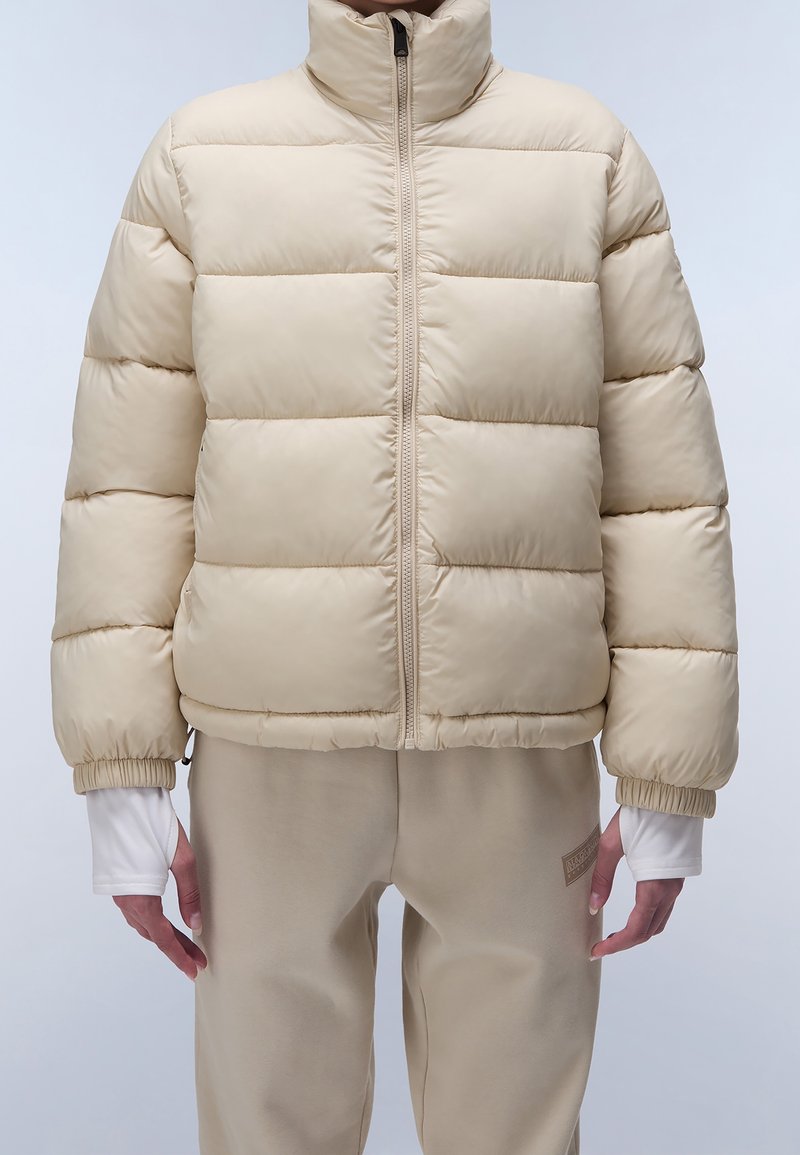 Beige pufferjacka med hög krage, horisontell sömnad, elastiska muddar och dragkedja framtill. Bärs över matchande beige byxor.