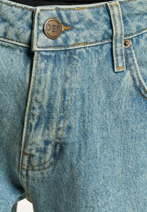 Lyseblå denim jeans med falmet struktur, orange syninger og en metalknap med "DEF" graveret. Side lomme synlig.