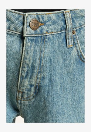 Világoskék farmernadrág, elhalványodott textúrával, narancssárga varrással és "DEF" felirattal gravírozott fém gombbal. Oldalsó zseb látható.