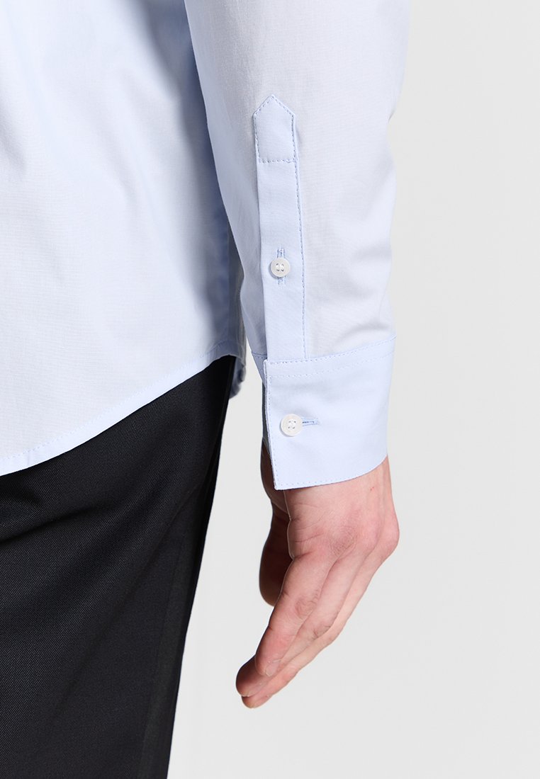 Himmelblaues Langarmhemd mit Knopfmanschetten-Detail, featuring einer glatten Textur und einem spitzen Kragen. Getragen mit schwarzen Hosen.