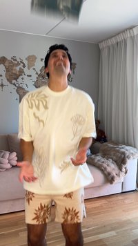 Weißes Kurzarm-T-Shirt mit beigen Blumen- und Wellenmustern, kombiniert mit hellbeigen Shorts mit Sonnenmotiven, in einem gemütlichen Innenraum.