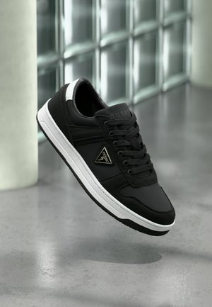 Μαύρο αθλητικό παπούτσι Guess με λευκή σόλα και τρίγωνο λογότυπο στο πλάι, αιωρούμενο πάνω από λείο γκρι πάτωμα με φόντο τοίχο από γυάλινα τούβλα.