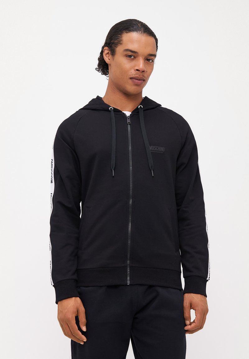 Črni zip-up hoodie z zadrgo spredaj, vrvicami in kontrastnimi stranskimi črtami, ki prikazujejo besedilo logotipa. Mehka tkanina, rebrasti manšeti in rob.