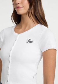 Bílý žebrovaný top s krátkým rukávem, kulatým výstřihem, zapínáním na knoflíky a vyšitým černým logem "Guess Jeans" na levé části hrudi.
