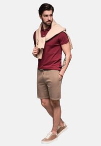 Shorts beige, polo shirt bourgogne, et pull beige drapé sur les épaules. Baskets en mélange de beige et de blanc. Tenue décontractée.