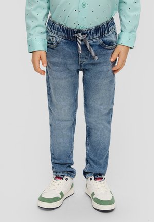 Jeans in denim blu con vita elastica e cordoncino a righe, dotati di tasche frontali e vestibilità slim. Sneakers bianche con accenti verdi.