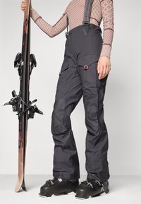 Ski pants in dark gray fabric with adjustable straps, side pockets, and reinforced knees. Paired with black ski boots and skis.