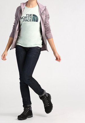 Hellviolette Kapuzenjacke mit Reißverschluss, mintgrünes T-Shirt mit Logo, dunkle Jeans und schwarze Wanderschuhe. Strukturierter Stoff und lockere Passform.