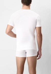 Uomo con una camicia bianca a maniche corte aderente e boxer bianchi, in piedi contro uno sfondo chiaro e uniforme, di spalle.