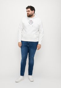 Felpa bianca con cappuccio caratterizzata da un logo centrale, tasca a marsupio e coulisse. Abbinata a jeans blu scuro e sneakers bianche. Design semplice e casual.