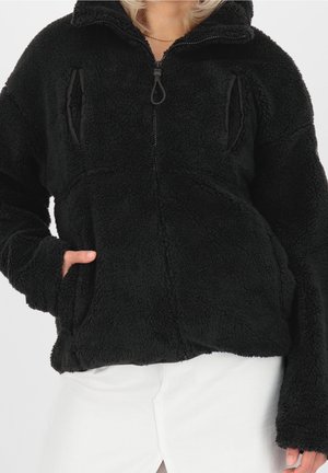 Personne portant une veste noire duveteuse à fermeture éclair avec col haut et poches latérales, portée avec un pantalon blanc, sur un fond uni.