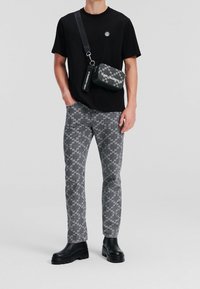 Camiseta negra, vaqueros grises con patrón y botas gruesas negras. El bolso cruzado presenta un estampado gráfico y acentos de logo.